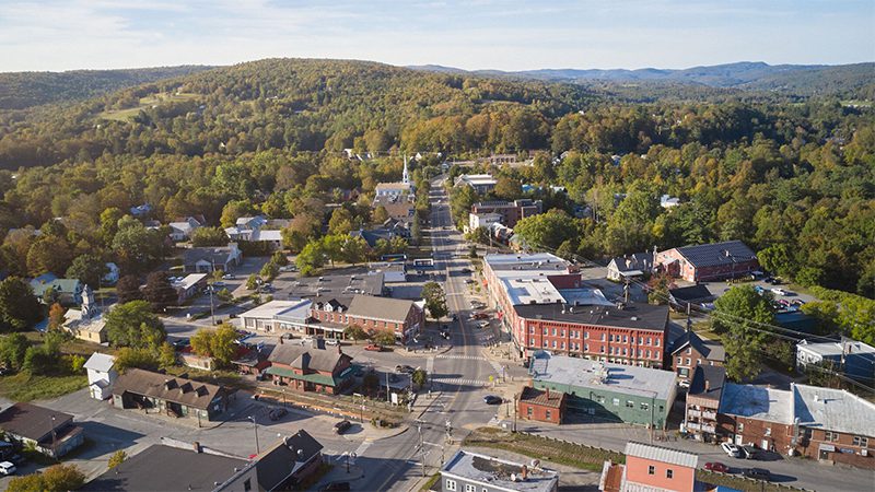 An aerial view shows a main street running through a small valley town, surrounded by dense green forests and rolling hills.