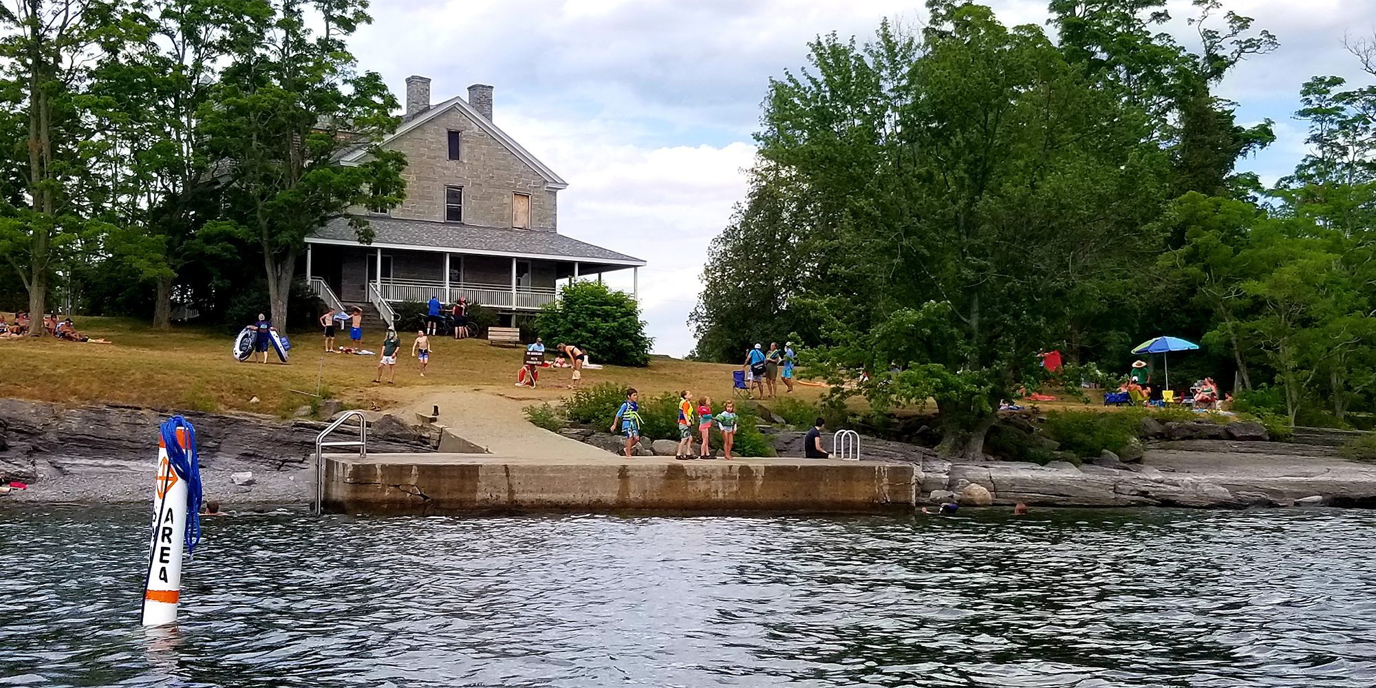 People gather on a grassy lawn outside a historic building in the summer on a lake.