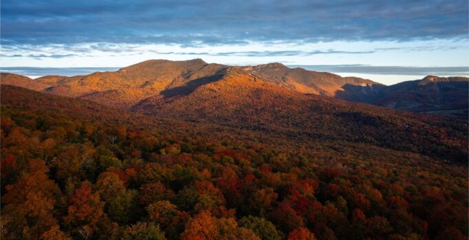 Seen from a distance, a mountain peak under cloudy skies covered in red, orange, and yellow leaves, creating an impressive display.