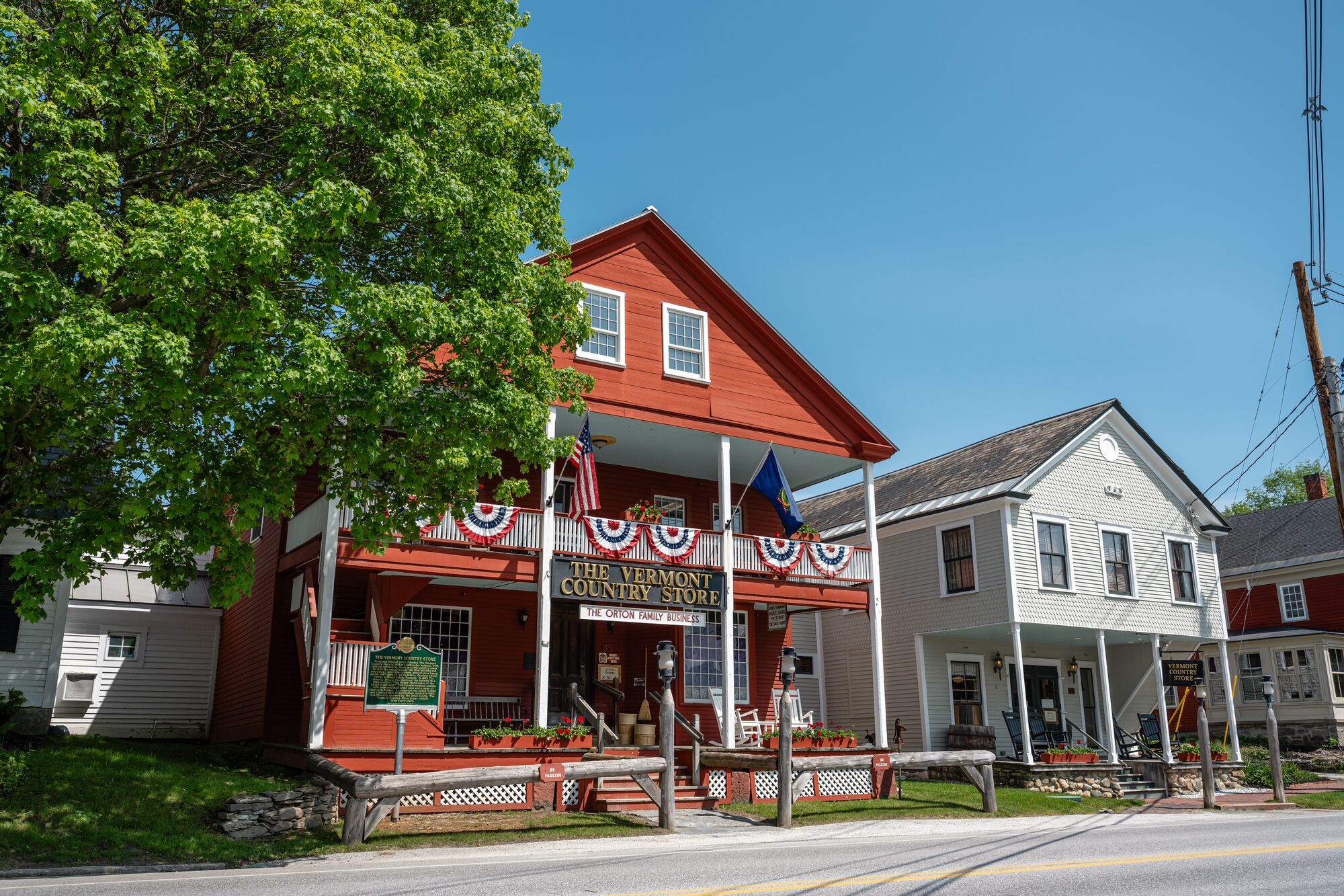 The large red wooden storefront of The Vermont Country Store, decorated with patriotic flags and bunting, stands next to a historic grey building under a clear blue sky.