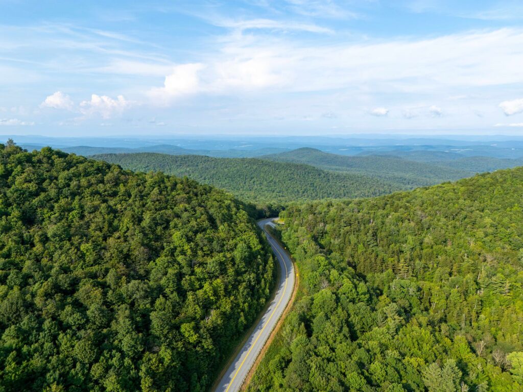 An aerial view captures a winding road cutting through a vast expanse of lush, green forested mountains under a partly cloudy sky.