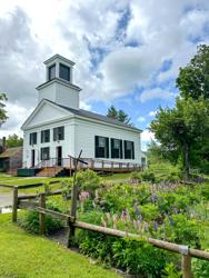 A historic white wooden building with a tiered bell tower stands on a grassy lawn behind a rustic wooden fence and garden under a partly cloudy sky.