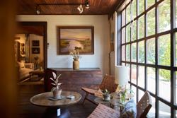 A sunlit, rustic sitting area features woven wooden chairs, a low coffee table with potted plants, and a landscape painting hanging above a wooden cabinet next to a large grid window.