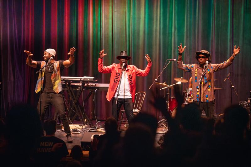 Three performers sing and dance on a brightly lit stage in front of a colorful, vertically striped curtain as an audience watches.