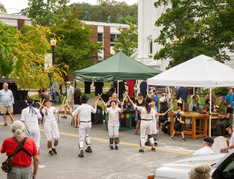 A group of dancers dressed in white perform a traditional Morris dance with sticks on a paved street at an outdoor festival with event tents and spectators in the background.