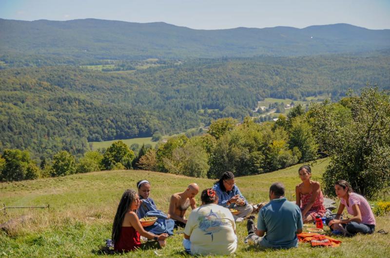 A group of people sit in a circle on a grassy mountain meadow enjoying an outdoor picnic together.