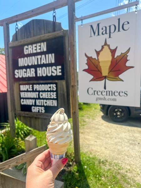 A hand holding a creemee in a cone in front of outdoor signs for the Green Mountain Sugar House.