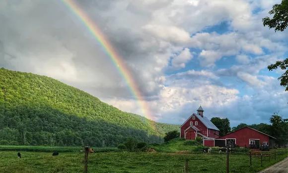 A bright, colorful rainbow arcs across a cloudy sky over a lush green hillside and a red farm house with matching barns.