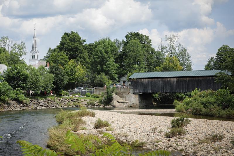 A wooden covered bridge stretches across a river in the summer. A church spire is visible on the left side.