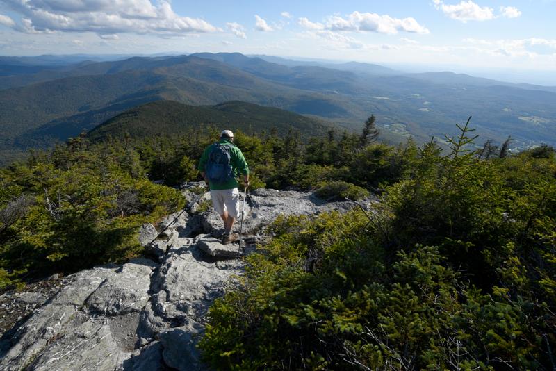 A hiker stands on a rocky mountain summit looking out over a vast mountain valley under a blue sky.