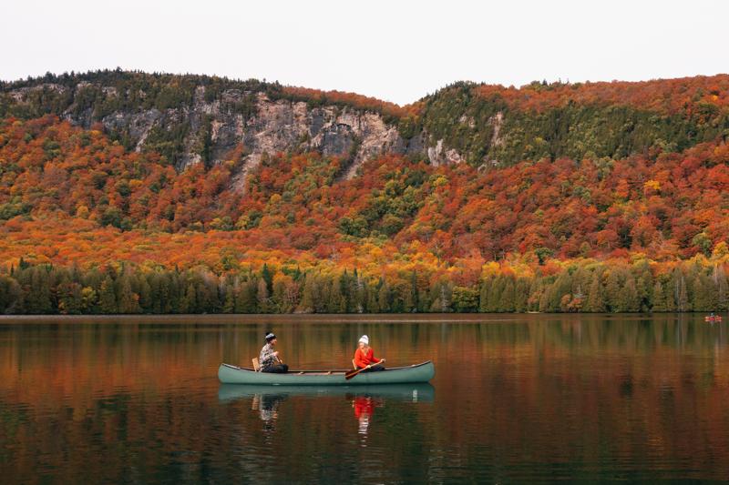 Two people in a canoe in front of a mountain covered with trees showing red, orange, and yellow leaves. Overlaid is a graphic of Vermont with red, orange, yellow, and green visible and regions outlined.