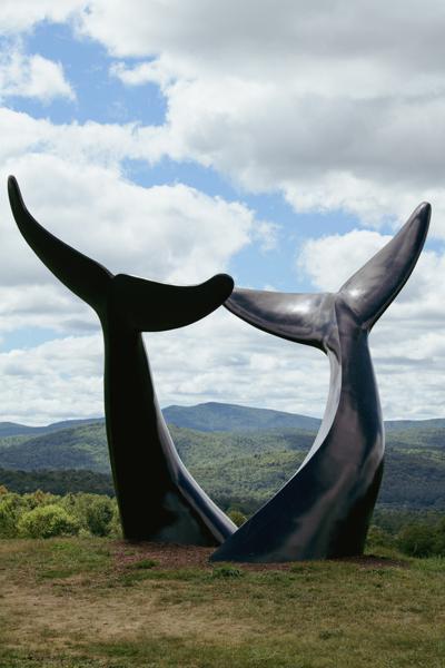 A large outdoor sculpture of two black whale tails curving toward each other sits on a grassy hill overlooking distant mountains under a cloudy sky.
