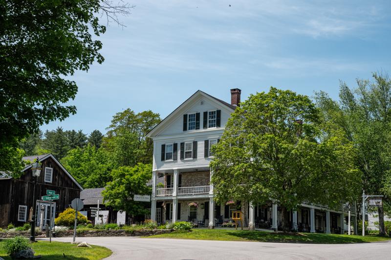 A historic, multi-story white building with black shutters and a covered two-tier porch stands surrounded by lush green trees under a bright blue sky.
