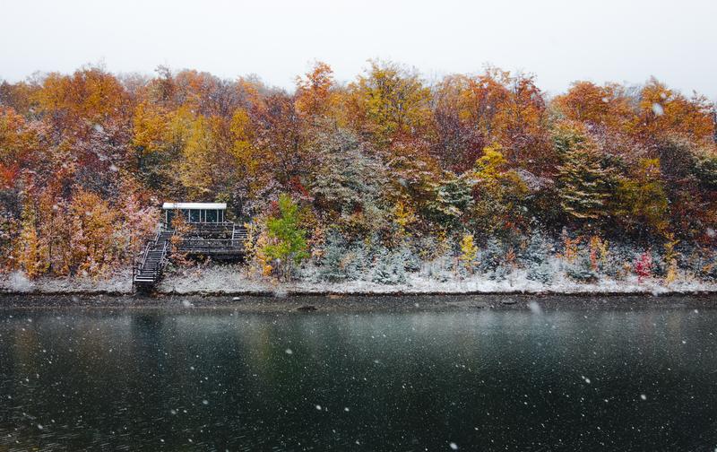 Next to a lake, a forest still shows red, orange, and yellow leaves on some trees, though some have fallen. Snow is falling and is sticking to the forest floor. Overlaid is a graphic of Vermont with regions outlined and most of the state beige, with red and orange visible on the left.
