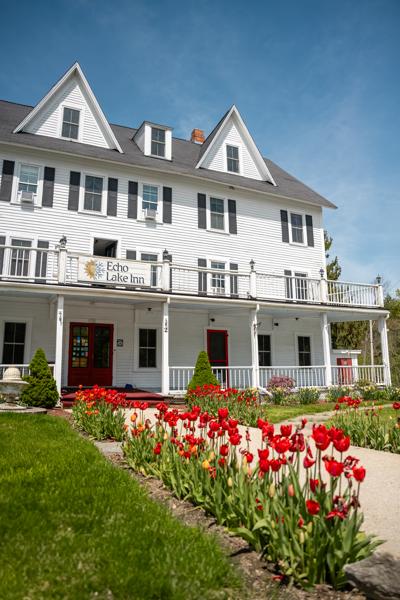 Bright red tulips line a walkway leading up to a historic, multi-story white inn with a large covered front porch under a clear blue sky.