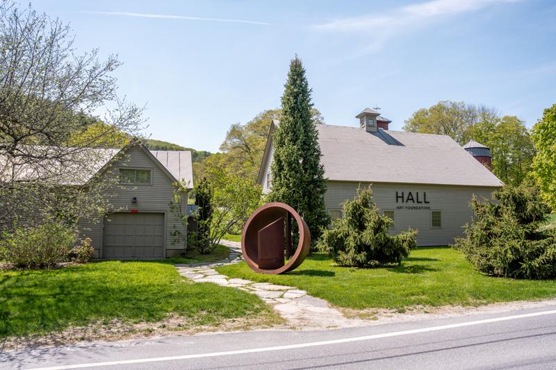 A large circular outdoor sculpture sits on a grassy lawn between two grey rustic buildings under a clear blue sky.