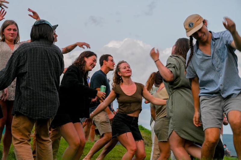 A group of people dance and laugh together outdoors on a grassy hill.