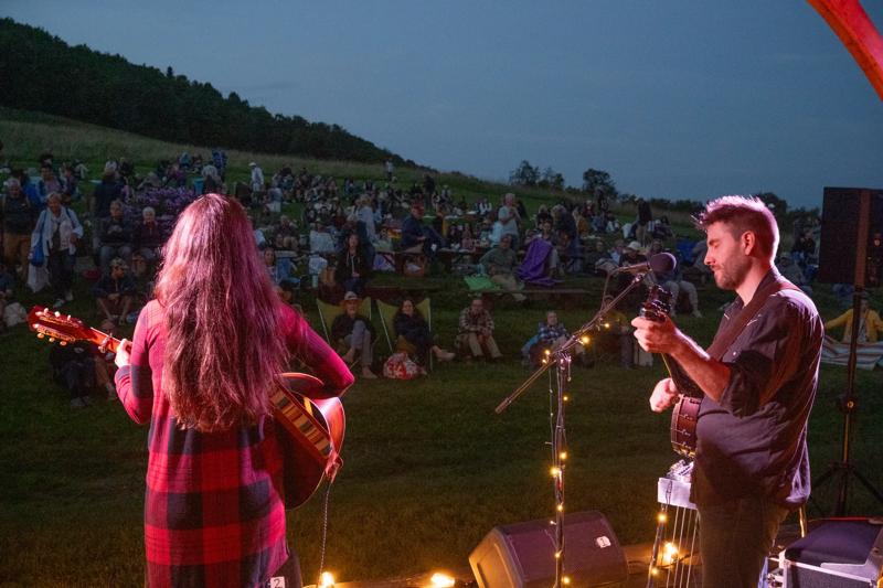 Musicians perform on an outdoor stage at dusk before a large crowd seated on a grassy hillside.