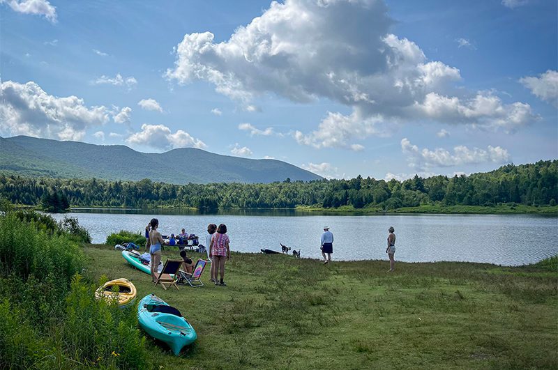 People gather on a grassy shoreline next to colorful kayaks parked by a calm lake with mountains in the background.