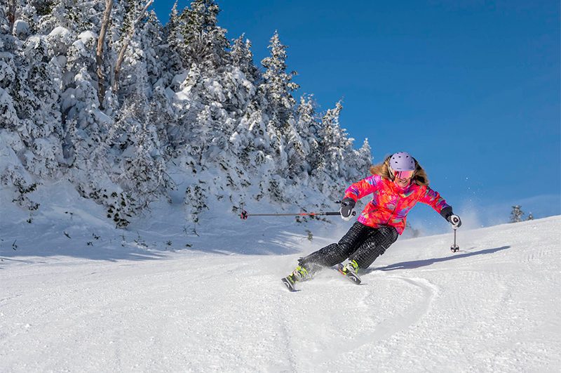 A skier dressed in bright pink down-turns on a groomed ski slope next to snow-covered evergreen trees.