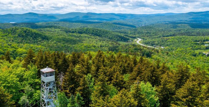 Seen from above, a fire tower in a dense forest with mountains visible around on a sunny summer day.