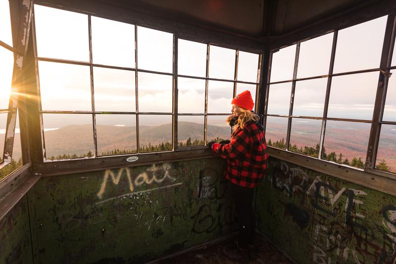 A person in a plaid jacket and beanie hat takes in the view at the top of a fire tower in the fall at sunset.