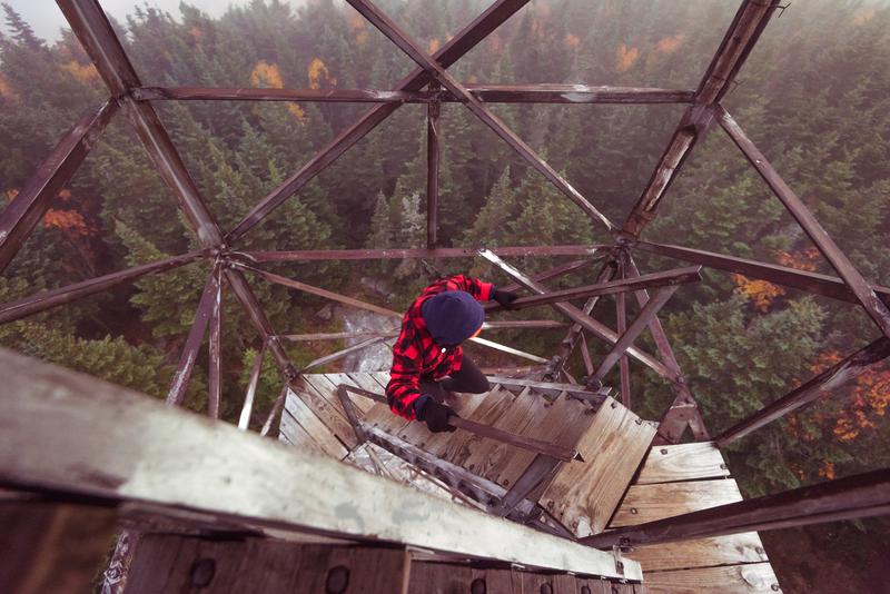 A high-angle, looking-down view of a woman climbing the wooden and metal stairs of the Bald Mountain fire tower.