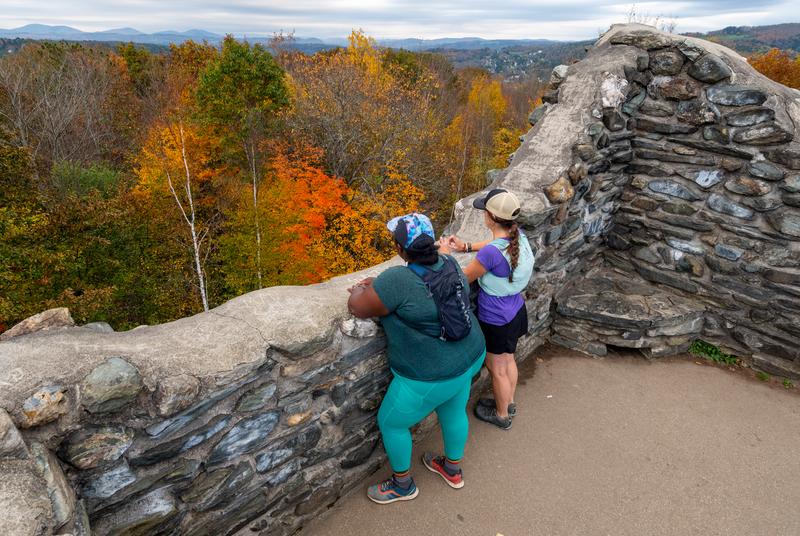 Two people take in the view from the top of a stone tower, looking over trees with leaves in red, orange, and yellow in the fall.