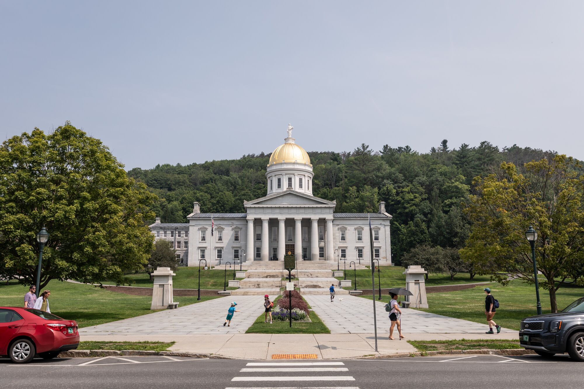A wide front view of the Vermont State House in Montpelier, featuring its iconic golden dome and white columns, with a paved walkway leading up to the entrance through a green lawn where people are walking.