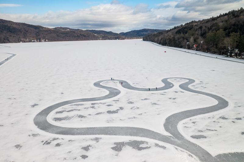 An aerial view of a wide, snow-covered frozen lake surrounded by forested hills under a cloudy sky. A winding ice skating path is cleared through the snow, forming a wide, looping circuit with several people skating along it.