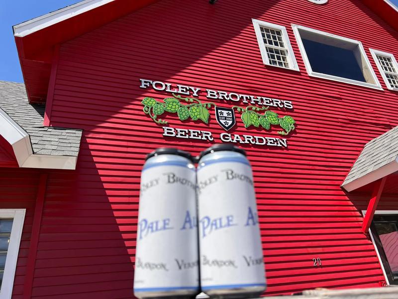 A close-up view of two silver cans of Foley Brothers Pale Ale held in the foreground, with the vibrant red siding and sign of the Foley Brothers Beer Garden barn visible in the background.