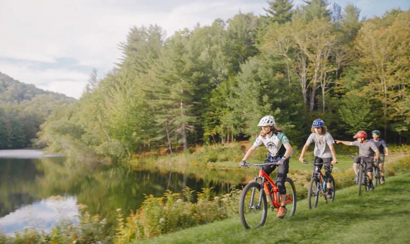 Four bicyclists wearing helmets ride mountain bikes in a single-file line along a grassy path beside a calm lake surrounded by dense green forest.