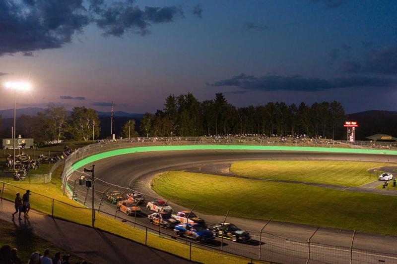 A high-angle view of race cars speeding around a curved asphalt track illuminated by stadium lights at dusk, with packed grandstands and dark green trees lining the outer edge under a fading twilight sky.