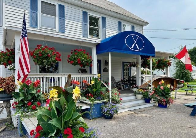 The exterior porch of a two-story white building decorated with vibrant red, yellow, and purple potted flowers, hanging baskets, an American flag, a Canadian flag, and a prominent blue awning featuring a white logo with crossed paddles.
