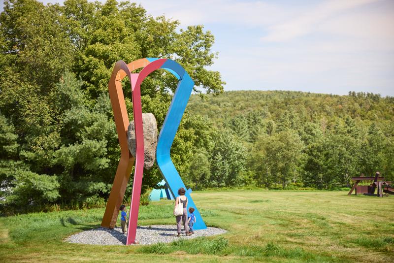 An adult and two young children stand on a grassy field looking up at a large outdoor sculpture featuring curved, colorful metal beams in shades of orange, red, and blue that hold a massive boulder suspended in the center, with a forested hill in the background.