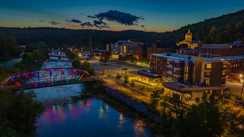 Aerial drone view of downtown Montpelier, Vermont at dusk, showing illuminated bridges crossing the river and warm lights across the city.