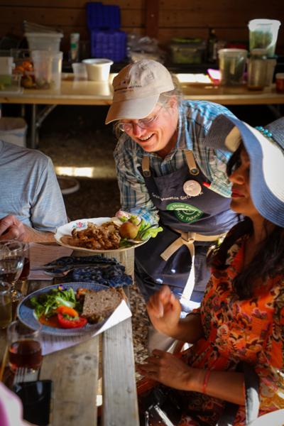 A waiter wearing a tan cap and a dark apron smiles as they serve plates of fresh salad and cooked dishes to guests seated at a rustic wooden picnic table during an outdoor dining event.