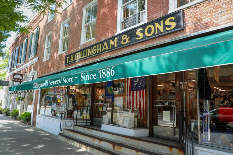 Exterior view of F.H. Gillingham & Sons general store with a dark sign above a long green awning and large glass storefront windows.