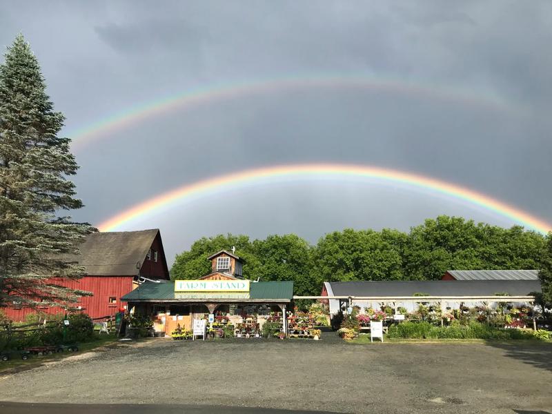 A vivid double rainbow arches across a stormy sky over a farm stand and red barn.