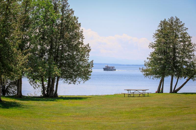 A wooden picnic table sits on a wide green grassy lawn framed by tall evergreen trees, looking out over a calm blue lake where a ferry boat travels in the distance under a hazy sky.