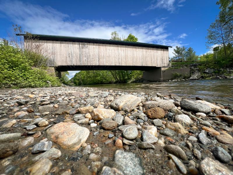 A low-angle view of a rocky, pebble-filled riverbank in the foreground, with a historic wooden covered bridge spanning across a shallow, flowing river under a bright blue sky with white clouds.