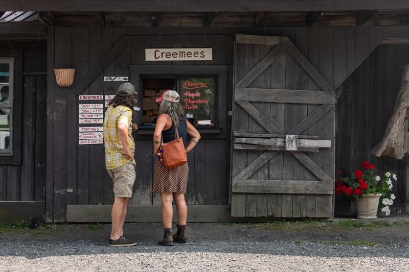 Two people stand outside a rustic, dark wood farm stand window with a sign overhead that reads 