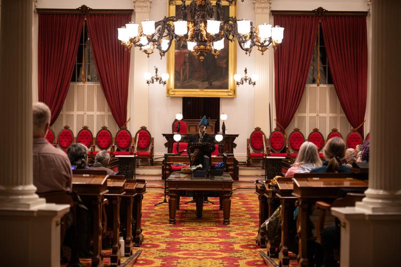 An interior view of a historic legislative chamber, featuring rows of ornate red chairs, heavy red curtains, and a large central chandelier, with an audience seated in the foreground listening to a person playing an instrument at a podium.