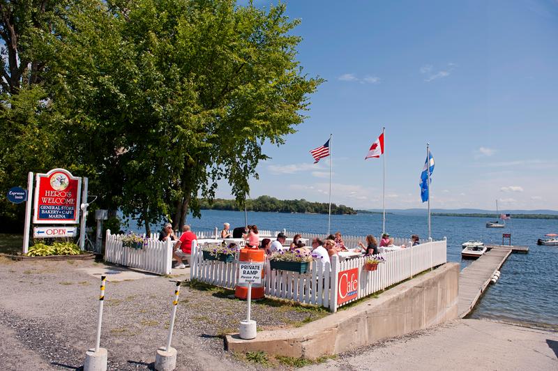 Customers dine at an outdoor lakeside patio enclosed by a white picket fence at Hero's Welcome, with a boat dock, flying flags, and a wide blue lake visible under a clear sky.