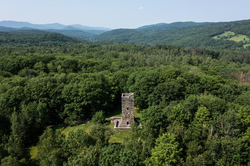 An aerial view of a tall, historic stone tower surrounded by a dense green forest, with rolling hills and mountains stretching into the distance under a hazy blue sky.