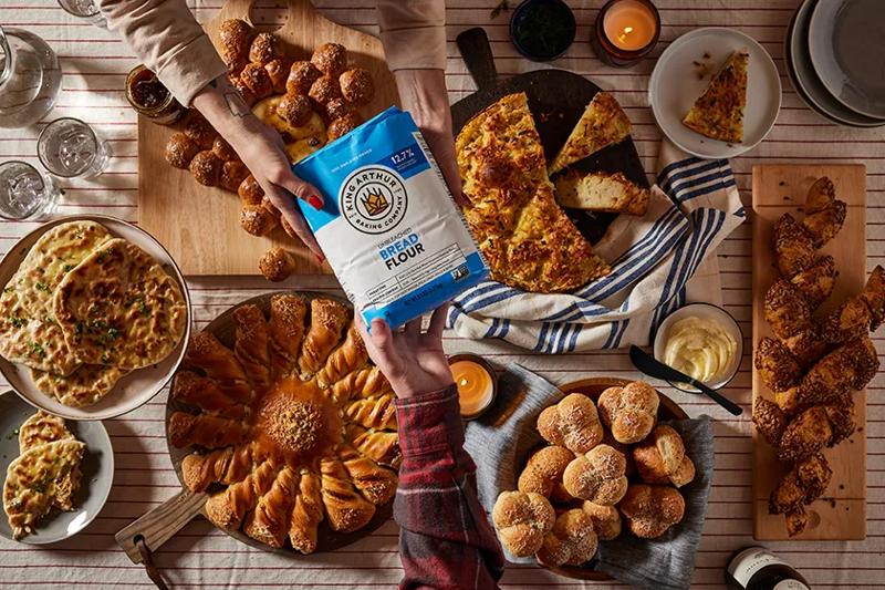 An overhead view of two hands passing a bag of King Arthur Bread Flour across a table filled with various freshly baked breads, rolls, and pastries.