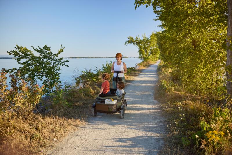A person pushes two young children in a stroller wagon along a sunny dirt recreation path lined with green trees, with a calm lake visible on the left.