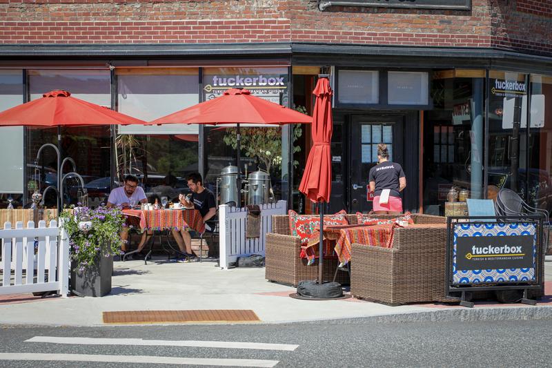 People dine at outdoor patio tables shaded by large red umbrellas in front of a brick restaurant building with a 