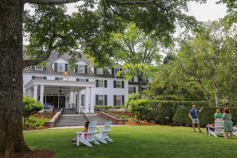 A large, historic white building with dark shutters and a grand columned entrance sits behind a lush green lawn, where several people relax and converse in Adirondack chairs under the shade of mature trees.