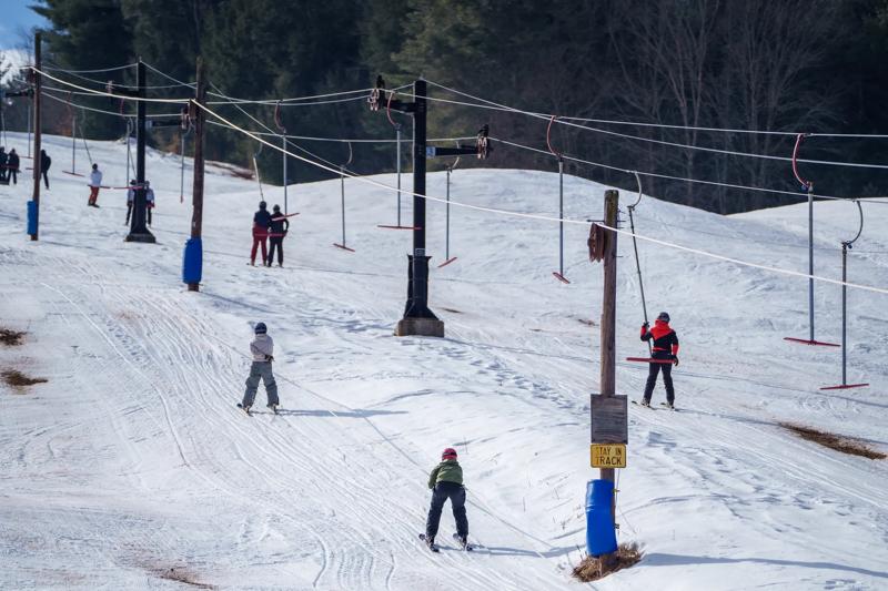 Skiers and snowboarders ride surface lifts up a snowy slope flanked by evergreen trees on a clear day.